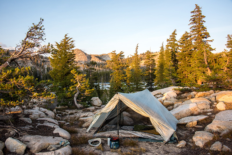 Tent and simple camp on decomposed granite with surrounded by boulders and dwarf trees, slight glimpse of Mattie Lake in background.